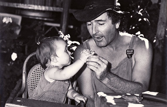 A baby girl sitting in a high chair reaching to her smiling father who is sitting at a table beside her.