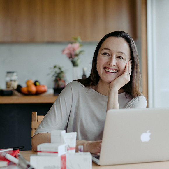 A woman sitting in front of a laptop and skin care products smiling.