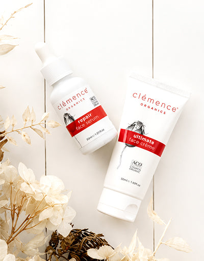 Two organic skincare products from Clémence Organics developed to treat ageing & mature skin: a white bottle of Repair Face Serum and a white tube of Ultimate Face Crème, both with red labels, arranged on a white wood background with dried flowers.