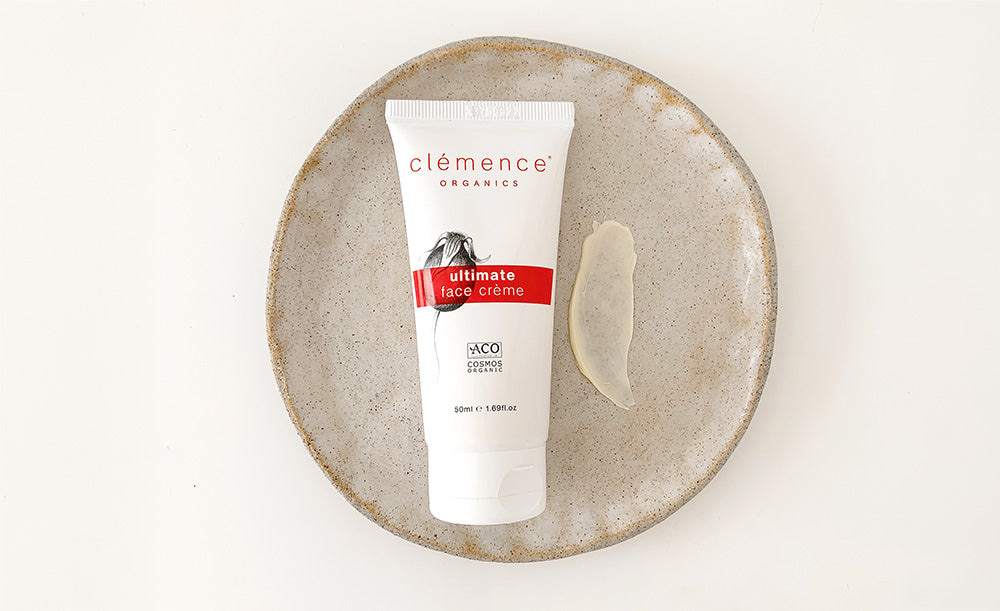 A white tube of Clémence Organics Ultimate Face Crème sits on a round, speckled beige ceramic plate next to a smear of pale cream, photographed from above on a white background.