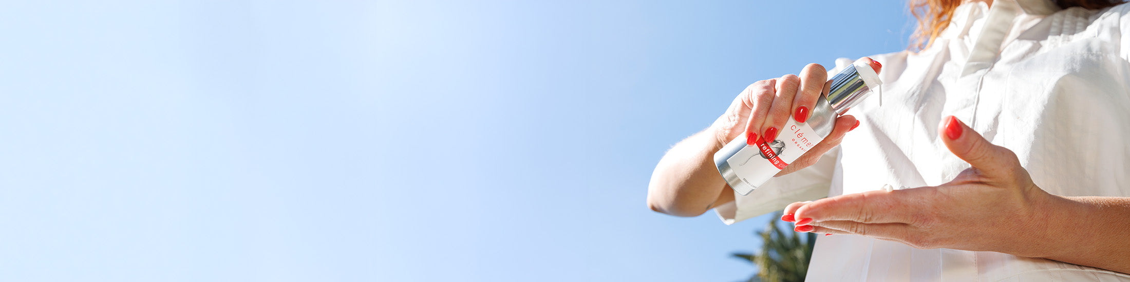 A close up lifestyle shot of a person applying cleanser from bottle with a white cap onto their hand, set against a bright blue sky.