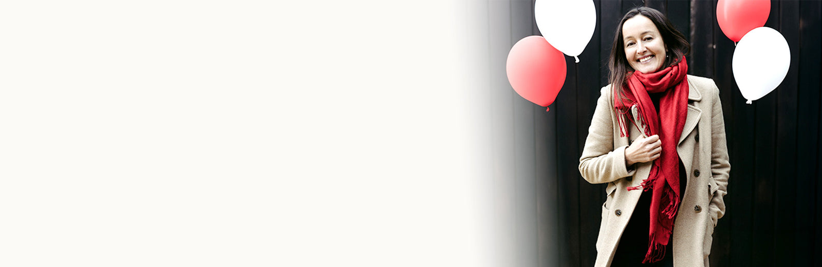 A woman wearing a beige coat and a bright red scarf stands against a dark background with red and white balloons floating near her.