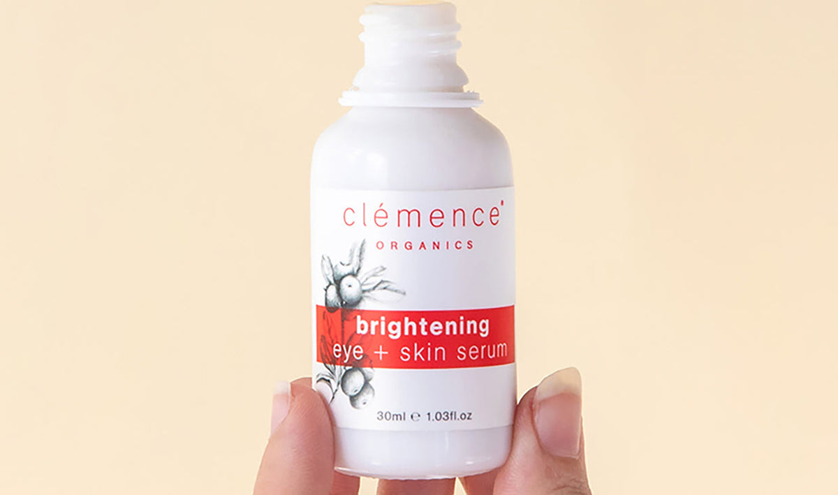 A hand holding a white bottle of clémence Organics Brightening Eye + Skin Serum against a solid pale beige background.