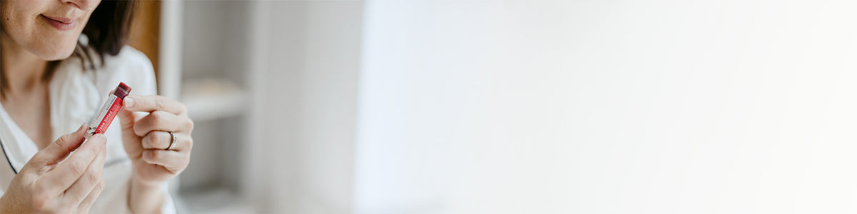 A woman holding a Clémence Organics Rose Tinted Lips tube, shown in a brightly lit indoor setting with a large blank white space on the right.