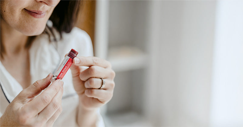 A close-up lifestyle shot of a woman holding a small red and white Clemence Organics Rose Tinted Lips tube, with a soft-focus indoor background.