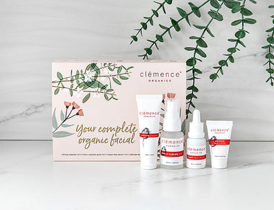 Clemence Organics 'Your complete organic facial' gift set displayed on a white marble surface with a white marble background and green eucalyptus branches. The set includes a large pink and white box with botanical illustrations, alongside four white skincare bottles and tubes with red and black labels.