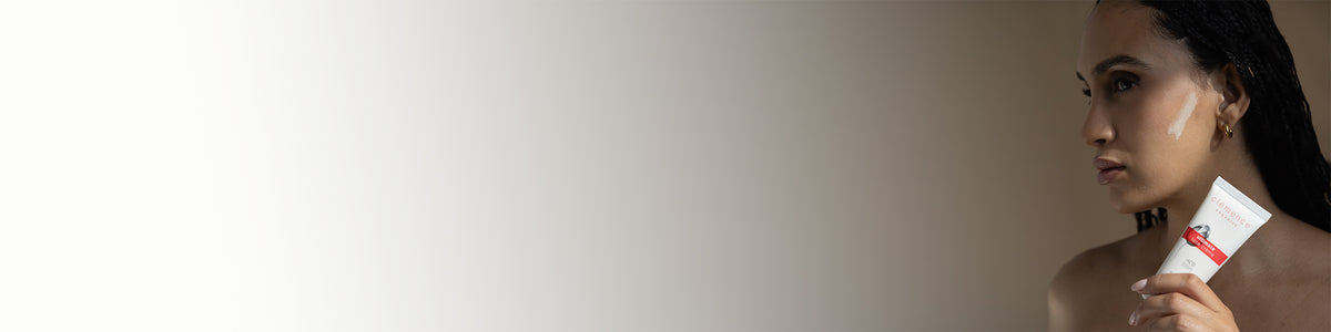 A woman in profile holding a white tube of Clémence Organics Ultimate Face Creme against a plain beige studio background.