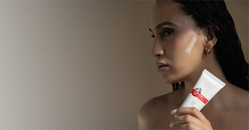 A woman in profile holding a white tube of Clémence Organics Ultimate Face Creme against a plain beige studio background.