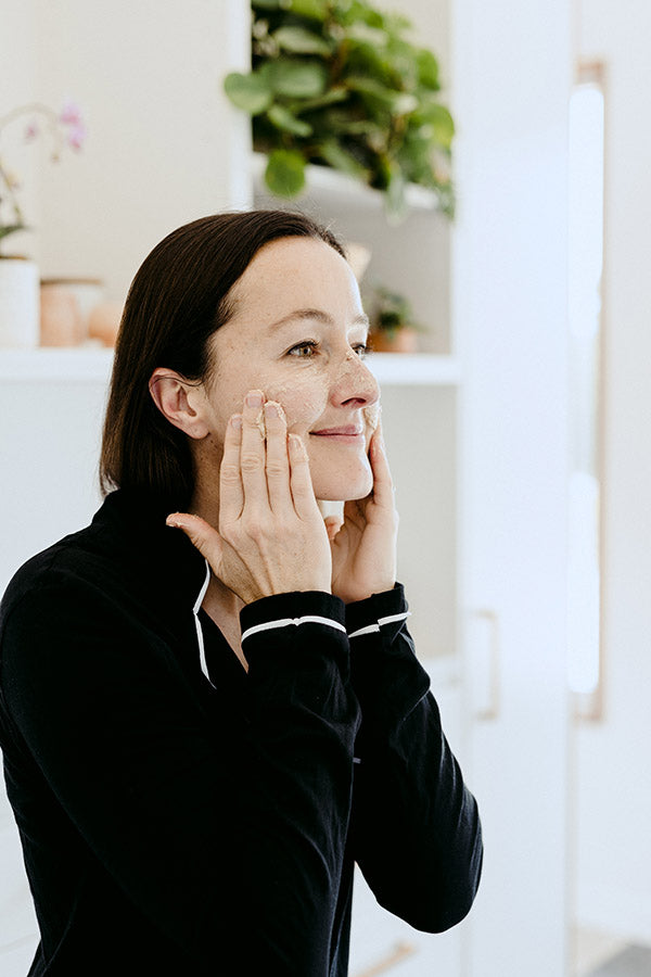 A woman applying an exfoliant & mask skin care product to her face.