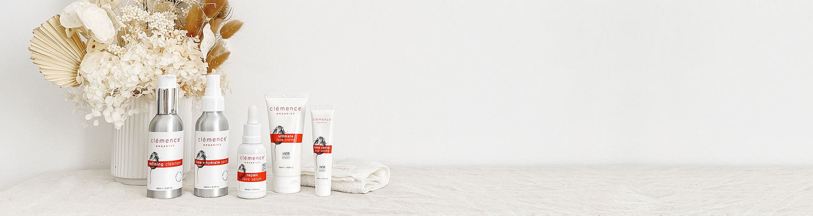 Five organic skin care products and a face cloth on a table. Behind the skin care products is a vase with flowers.