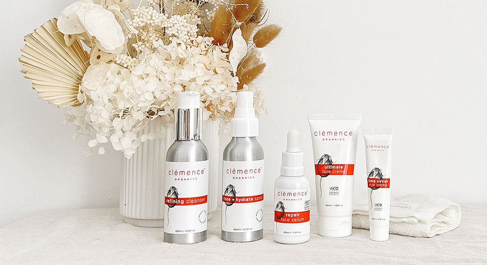 A set of five Clemence Organics skincare bottles and tubes arranged in front of a vase of dried flowers on a white surface against a white background.