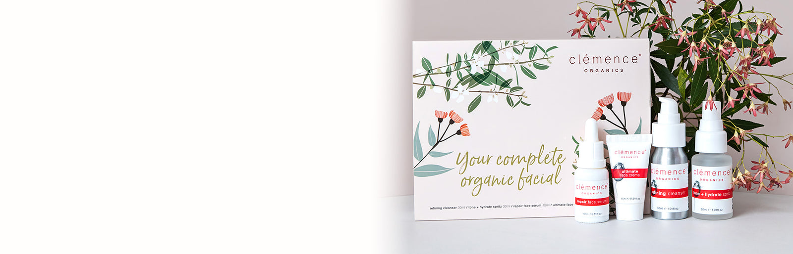 The Clémence Organics 'Mini' Face Essentials pack on a white surface with a soft pink background, including a floral box, a small bottle, and two dropper bottles with pink labels, next to pink flowers.