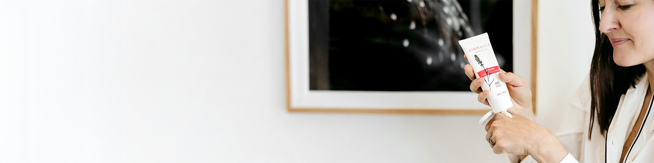 A woman on the right holds a white tube of Clemence Organics hand & body lotion with a red and black label, set against a bright white wall with a framed picture.