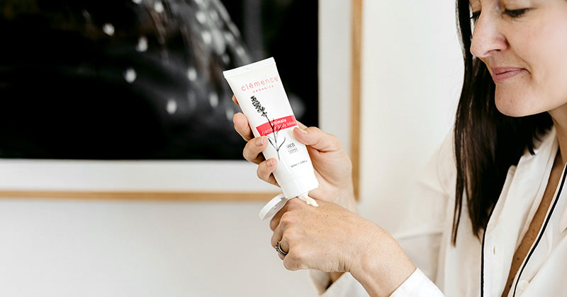 A woman in a white robe holds a white tube of Clemence Organics Ultimate Hand & Body Lotion in front of her face, with a framed black and white artwork on a light wall in the background.