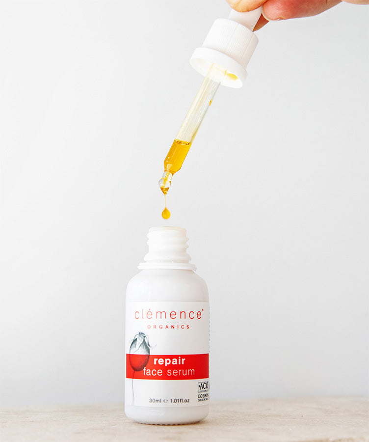 A white bottle of Clémence Organics repair face serum with a white dropper cap, shown with a hand holding the dropper above a single drop of golden-yellow serum about to drip into the bottle, on a light neutral background.