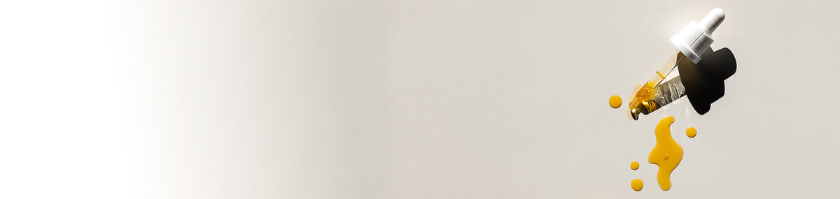 A single dropper-style cosmetic applicator with a white cap, shown on the right side of a plain light gray background with yellow liquid droplets splashing below it.