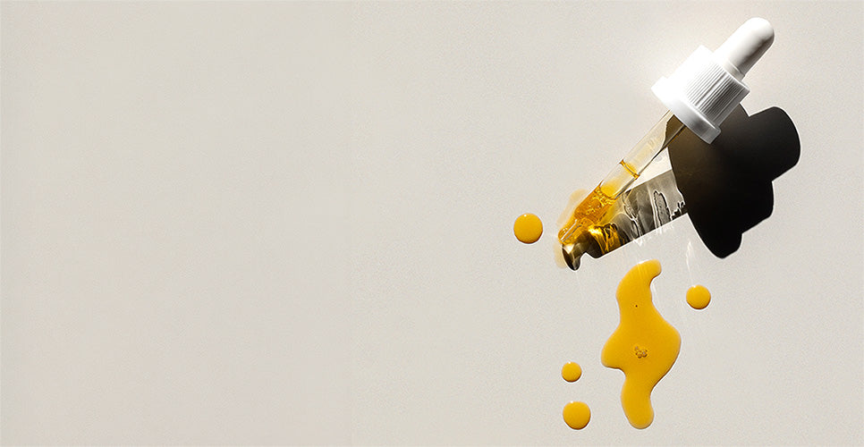 A close-up of a dropper bottle tipped over with golden yellow liquid dripping out, set against a plain light gray background.