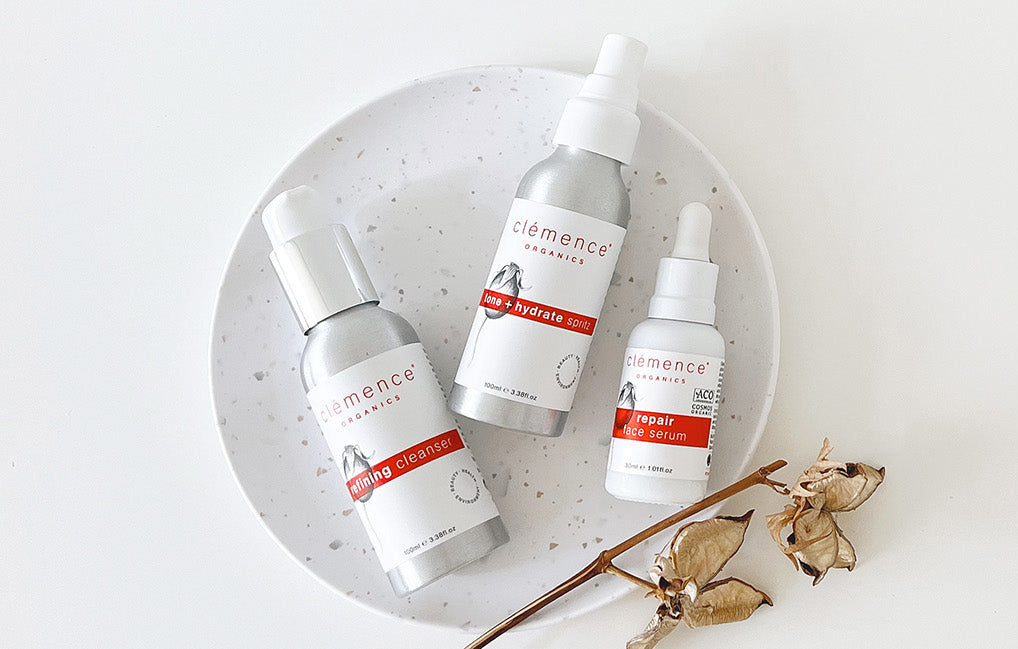 Three silver and white skincare bottles from Clémence Organics arranged on a speckled white plate with dried botanical stems, photographed from above on a white background.