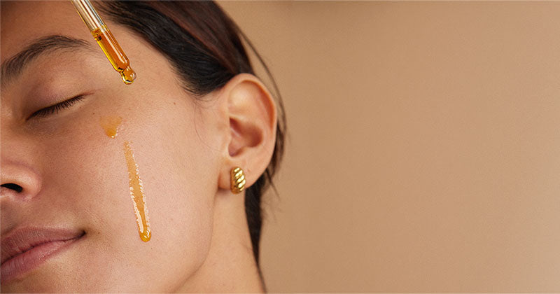 Close-up of a woman with closed eyes and gold ear cuff, with golden liquid droplets dripping from a dropper onto her cheek against a warm beige background.