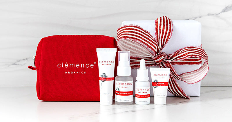 A Clémence Organics gift set featuring a red zippered pouch, four white skincare bottles and tubes with red and black labels, and a white gift box tied with a red and white striped ribbon, arranged on a white marble surface.