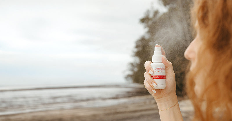 A woman standing on a beach holds and sprays a bottle of Clémence Organics Ultimate Biome Spritz on her face.